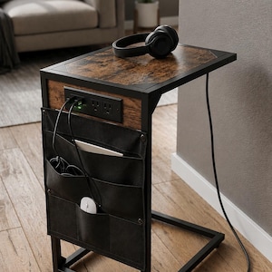 May include: A C-shaped side table with a dark wood grain top and a black metal frame. It features a built-in power strip with two electrical outlets and two USB ports, plus a black fabric storage pocket on the side containing a white computer mouse.