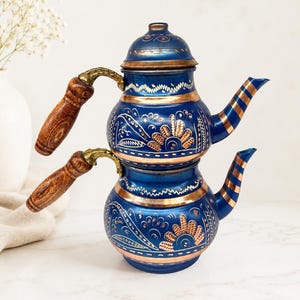 May include: A double-decker tea kettle crafted from copper with a vibrant blue finish. The exterior features intricate hand-engraved floral patterns revealing the copper base. It includes two wooden handles and two spouts with striped accents.