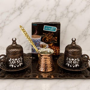 May include: A Turkish coffee set featuring two dark bronze-toned cups with ornate cut-out patterns and matching lids, a hammered copper coffee pot with a long handle, and a rectangular tray, all displayed with a box of Ali Baba Turkish Coffee.