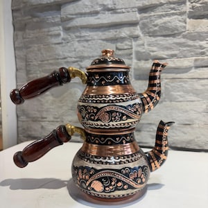 Può includere: Una teiera decorativa in rame e nera con un design a strati unico. Presenta motivi floreali ornati, manici in legno e un beccuccio con strisce nere e ramate. Il coperchio è nero con accenti ramati.
