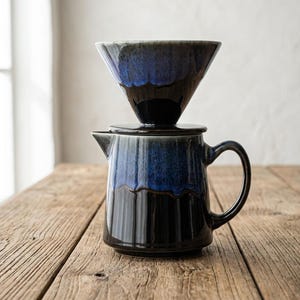 Puede incluir: Un juego de cafetera de cerámica que incluye un gotero cónico para café apoyado sobre una jarra a juego. Ambas piezas tienen un acabado brillante con una base negra profunda que se transforma en un esmalte azul vibrante en la parte superior.