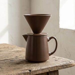 Op de afbeelding: Een matbruine keramische koffiezetset bestaande uit een conische filterhouder die stevig op een bijpassende cilindrische koffiekan met een zijhandvat en een kleine schenktuit rust voor het zetten van koffie.