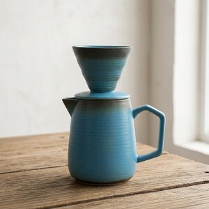 Può includere: Un set per caffè pour-over in ceramica composto da un portafiltro conico blu brillante appoggiato su una caraffa blu abbinata. Entrambi i pezzi presentano una superficie testurizzata e scanalata con una sfumatura grigio scuro sui bordi.