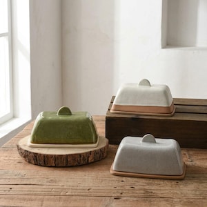 Puede incluir: Tres mantequilleras rectangulares con tapa, cada una de un color distinto: una verde oliva, una blanco crema y una gris claro moteado. Cada mantequillera tiene una base y una tapa a juego con un pequeño asa semicircular en la parte superior.