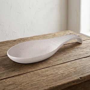May include: A white ceramic spoon rest with a speckled finish featuring small black dots across the surface. The spoon rest has an elongated oval bowl shape and a curved handle extension that slopes downward at the end for stability on surfaces.
