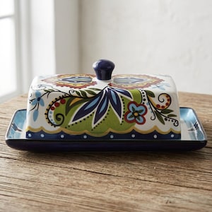 Puede incluir: Una mantequillera rectangular de cerámica con tapa que presenta un vibrante patrón floral y de cachemira multicolor en tonos verde, azul, rojo y amarillo. La base es de color azul oscuro sólido y la tapa tiene un pequeño pomo redondeado.