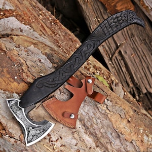 May include: A hand axe with a dark, carved wooden handle featuring intricate knotwork and a bird-shaped pommel. The metal blade displays detailed etched engravings of a bird and Celtic patterns. A tan leather sheath with metal snaps covers the blade.