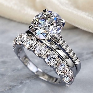 Peut inclure: Une bague en argent avec une grande pierre centrale ronde sertie de griffes, accompagnée d'une alliance assortie. Les deux pièces sont ornées de petites pierres rondes le long des anneaux, avec le poinçon 925 visible à l'intérieur.