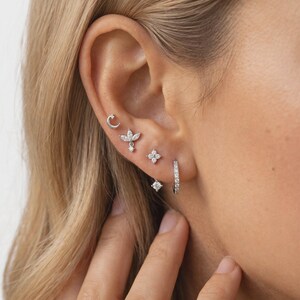 May include: A set of silver-toned earrings worn in a single ear, featuring a small crescent moon stud, a leaf-shaped charm stud, a four-petaled flower stud, a small dangling crystal stud, and a thin hoop earring encrusted with tiny clear stones.