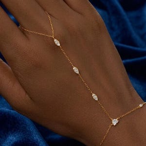 May include: A gold hand chain bracelet featuring a delicate fine link chain adorned with multiple marquise-cut clear stones. The design includes a ring component connected to the wrist chain by a central strand, creating a continuous jewelry piece.