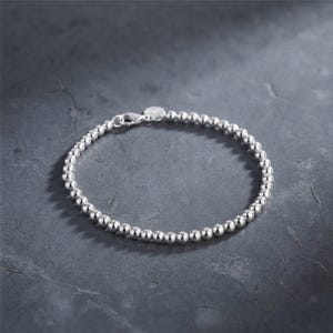 May include: A polished silver bead bracelet featuring a continuous strand of small, uniform spherical beads. The bracelet is secured with a silver lobster claw clasp and includes a small, round metal tag attached near the closure mechanism.