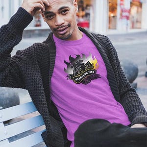May include: A person wearing a purple t-shirt with the text "HEARTS ON FIRE" in a banner design. The shirt has a graphic of flames in black, gray, and yellow. The person is also wearing a dark gray cardigan and black pants.
