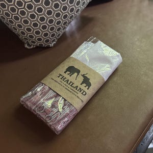 May include: A folded textile wrapped in clear plastic with a tan paper band featuring two black elephant silhouettes, the text THAILAND, 100% SILK & CASHMERE, and THE BEST GIFTS TO IMPRESS YOUR BELOVED. The fabric displays a maroon and white pattern.