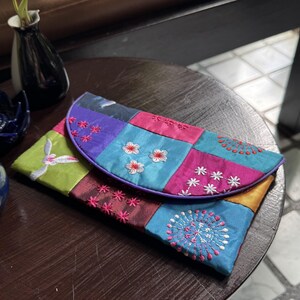 Op de afbeelding: Een rechthoekige clutch met een patchworkontwerp van zijdeachtige stof in levendige tinten groen, blauw, paars en roze. De flap is versierd met ingewikkeld wit en roze bloemenborduurwerk en cirkelvormige patronen over het gehele oppervlak.