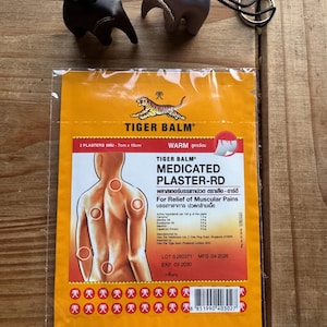 May include: Two small elephant-shaped leather keychains in dark brown and light gray, positioned above a rectangular orange package of Tiger Balm Medicated Plaster-RD. The package features a tiger logo, text, and a diagram of a human back with red circles.