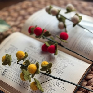 May include: Three decorative stems featuring felted wool berries in yellow, red, and off-white. Each stem includes small green felt leaves attached to thin, dark wire branches. The berries are round and textured, arranged along the length of the stems.