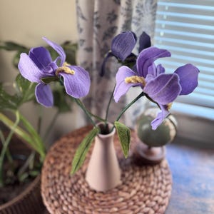 May include: Two artificial purple iris flowers with yellow centers and green leaves are arranged in a slender, matte light pink vase. The flowers feature textured petals and thin stems, creating a botanical display with a soft, muted color palette.