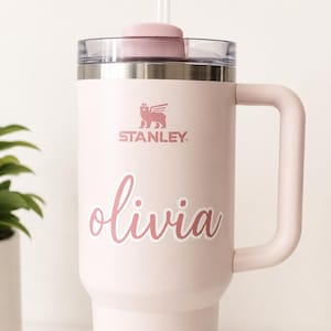 Peut inclure: Une tasse Stanley rose clair avec une poignée, un couvercle transparent et une paille rose. La tasse porte le nom "Olivia" en écriture cursive et le logo Stanley. La tasse est conçue pour garder les boissons froides ou chaudes.