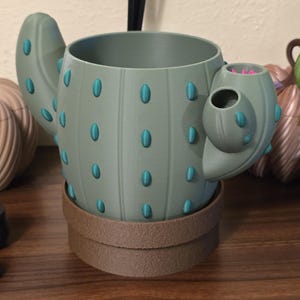 May include: A sage green cactus-shaped planter with a cylindrical body, featuring raised teal oval accents and two side arms. The planter sits inside a brown, textured circular base. The interior of the right arm shows a bright pink interior detail.