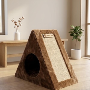 Puede incluir: Una casa triangular de felpa marrón para mascotas con un panel rascador de sisal texturizado en la parte frontal. Una pequeña placa rectangular de madera está fijada en la parte superior con el texto YOUR PET NAME y un icono de huella.
