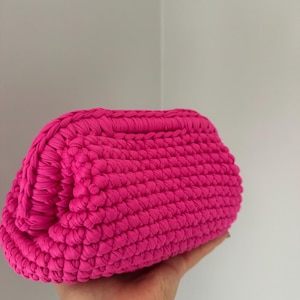 Könnte beinhalten: Eine handgefertigte Clutch-Tasche in leuchtendem Pink. Die Tasche besteht aus einem dicken, strukturierten Stoff, hat eine abgerundete Form und ein schlichtes, elegantes Design. Die Tasche wird in einer Hand vor einem schlichten weißen Hintergrund gehalten.