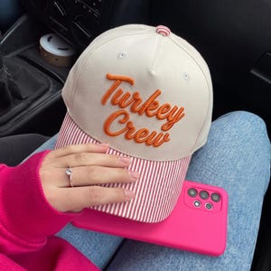 May include: A cream-colored baseball cap featuring the embroidered orange text "Turkey Crew" on the front panel. The cap has a red and white striped brim and a matching red and white striped button on the top center of the crown.