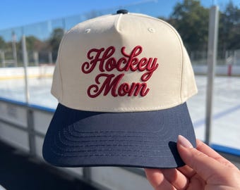 May include: A cream-colored baseball cap with a dark navy blue brim. The front of the cap features the text Hockey Mom embroidered in a dark red script font. The cap has a structured crown with a button on top and visible stitching on the brim.