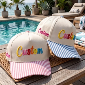 May include: Two cream-colored baseball caps with five-panel construction. Each cap features the word Custom embroidered in raised, multi-colored letters across the front. One cap has a pink and white striped brim, while the other has a blue and white striped brim.
