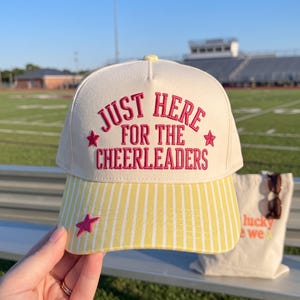 May include: A cream-colored baseball cap with a yellow and white vertical striped brim. The front features embroidered pink text that reads JUST HERE FOR THE CHEERLEADERS, flanked by two small pink stars, with an additional pink star on the brim.
