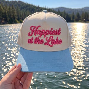 May include: A cream-colored baseball cap with a light blue brim. The front panel features the embroidered text "Happiest at the Lake" in a bold, cursive pink font. The cap has a structured crown and visible stitching along the brim.