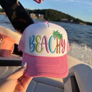 May include: A pink and white trucker hat featuring the word BEACHY embroidered in colorful script letters across the front panel, with two green palm trees integrated into the design, a solid pink brim, and a mesh back panel.
