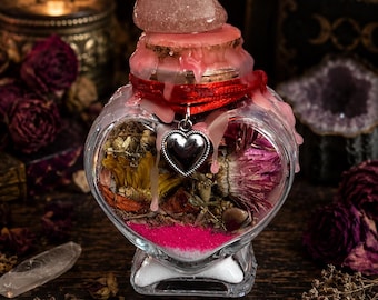 Può includere: Una bottiglia di vetro a forma di cuore riempita con fiori secchi, erbe e strati di sabbia rosa e bianca. Il tappo di sughero è sigillato con cera rosa, sormontato da una pietra rosa lucida e decorato con un nastro rosso e un ciondolo a cuore argentato.