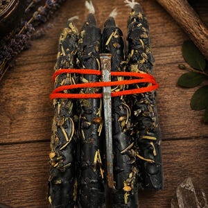 May include: A bundle of four black taper candles embedded with dried herbs and botanicals, secured together with bright orange twine and a single weathered, rusted metal nail placed vertically across the center of the candles.