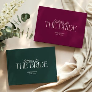 Peut inclure: Deux livres à couverture rigide rectangulaires avec le texte "letters to THE BRIDE" imprimé sur les couvertures. L'un est bordeaux foncé avec "ASHLEY ELISE Sept 26, 2026" et l'autre est vert forêt avec "MELISSA LYNN 12.06.2026" inscrit dessous.