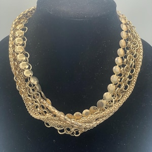 May include: A multi-strand gold-tone necklace featuring a variety of chain styles, including a strand of flat circular discs, a textured mesh chain, and several interlocking round link chains, all layered together to create a voluminous design.