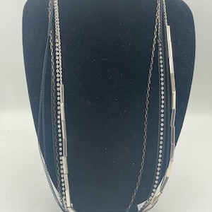 May include: A multi-strand necklace featuring a combination of thin black chains, a silver-toned chain with embedded clear rhinestones, and a chain accented with elongated silver-toned rectangular metal beads arranged in a layered design.