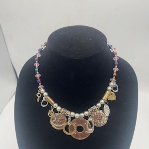 May include: A multi-strand necklace featuring a mix of pink and iridescent beads, silver-toned metallic spheres, and various dangling charms including a small key, circular metal discs, and textured geometric shapes in copper and silver tones.