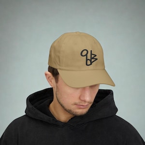May include: A tan baseball cap with a black abstract design on the front. The cap has a curved brim and a classic six-panel design. The cap is made of a soft material and is suitable for casual wear.