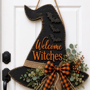 Puede incluir: Un letrero de puerta con forma de sombrero de bruja negro con acabado desgastado, que presenta tres pequeños murciélagos negros y el texto naranja "Welcome Witches". El letrero está decorado con un lazo a cuadros naranja y negro, arpillera, follaje verde y bayas naranjas.