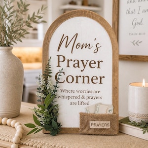 May include: An arched wooden sign with a white center and brown frame. The text reads: Mom's Prayer Corner, Where worries are whispered & prayers are lifted. It features a small burlap pocket labeled PRAYERS and a sprig of green artificial foliage.