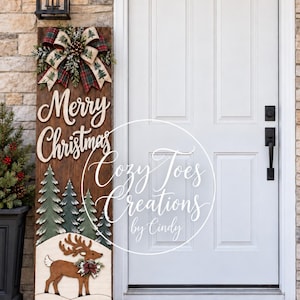 May include: A tall rectangular wooden porch sign featuring the text Merry Christmas in white script. The sign includes a decorative bow, painted green pine trees, and a brown reindeer figure with a small wreath around its neck.