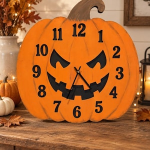 May include: A pumpkin-shaped wall clock with a bright orange exterior and a dark brown stem. The clock face features a black jack-o'-lantern cutout design for the eyes and mouth, with black numbers from 1 to 12 and two black clock hands.