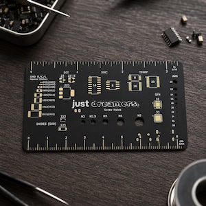 Op de afbeelding: Zwarte elektronische printplaat met gouden markeringen, inclusief SMD-, SOIC- en TSSOP-labels, samen met schroefgaten en een liniaal. De kaart heeft de tekst "just dreamers." en is omgeven door gereedschap en componenten.