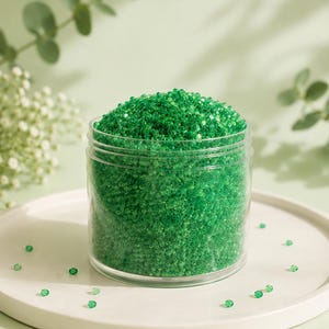 May include: A clear plastic jar filled with small, translucent emerald green seed beads. The jar is cylindrical with a flat base and a screw-top rim. Several individual green beads are scattered around the base of the container.