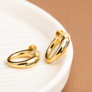 Op de afbeelding: Een paar goudkleurige oorringen met een gladde, gepolijste afwerking. Elke oorbel is voorzien van een klein schroefmechanisme en het woord Nirvana in zwarte letters gegraveerd langs de buitenrand van de gebogen metalen band.