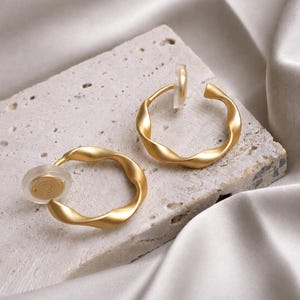 May include: A pair of gold-toned hoop earrings with a twisted, matte finish. Each earring features a clear, circular silicone clip-on backing attached to the hoop, designed for non-pierced ears, showcasing a smooth, metallic, and curved structure.
