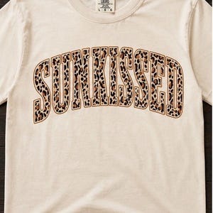 以下が含まれることがあります： 胸に「SUNKISSED」の文字がヒョウ柄で大きくプリントされた、クリーム色のTシャツ。クルーネックと半袖です。「Comfort Colors」のブランドがタグにプリントされています。