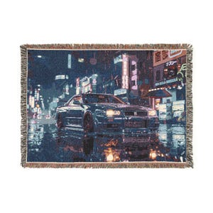 May include: Woven throw blanket depicting a dark blue sports car in a rainy city scene at night. The image includes neon signs and reflections on the wet pavement. The blanket has a fringed edge.