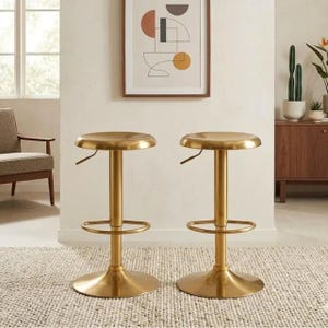 May include: A pair of gold-toned metal bar stools featuring a circular seat, a central cylindrical support column, a circular footrest, and a wide, flat, circular base. Each stool includes a small lever located just beneath the seat for height adjustment.
