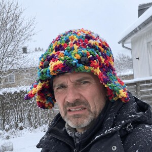 May include: A multi-coloured crochet hat featuring a vibrant spectrum of red, orange, yellow, green, blue, and purple yarn. The hat has a textured, bumpy surface and includes two large, round, black plastic eyes attached to the front section.